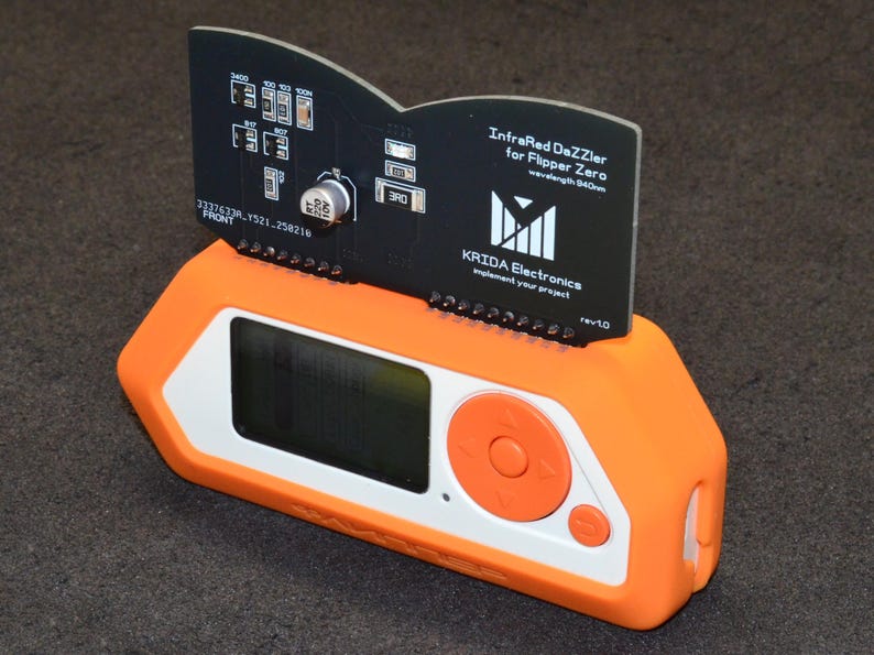 Puede incluir: Un dispositivo electr&oacute;nico naranja y blanco con una pantalla negra y botones. Una placa de circuito negro est&aacute; insertada en el dispositivo, mostrando el texto "InfraRed DaZZler for Flipper Zero" y el logotipo de KRIDA Electronics.