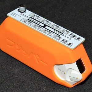 K&ouml;nnte beinhalten: Ein orangefarbenes elektronisches Ger&auml;t mit einer wei&szlig;en Leiterplatte oben. Die Leiterplatte hat mehrere kleine Lichter und einen silbernen Knopf. Das Ger&auml;t hat japanischen Text auf der Seite und einen wei&szlig;en Abschnitt mit Zahlen und Text.