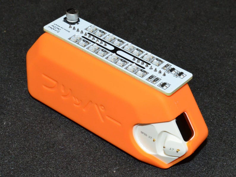 K&ouml;nnte beinhalten: Ein orangefarbenes elektronisches Ger&auml;t mit einer wei&szlig;en Leiterplatte oben. Die Leiterplatte hat mehrere kleine Lichter und einen silbernen Knopf. Das Ger&auml;t hat japanischen Text auf der Seite und einen wei&szlig;en Abschnitt mit Zahlen und Text.