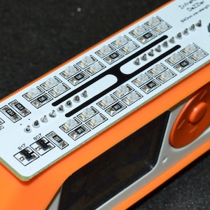 K&ouml;nnte beinhalten: Ein orange-wei&szlig;es elektronisches Ger&auml;t mit einer Leiterplatte mit zahlreichen kleinen Lichtern und Komponenten. Die Leiterplatte tr&auml;gt den Text "Infrared Dazzler" und "940nm wavelength". Das Ger&auml;t hat zwei orangefarbene Kn&ouml;pfe und eine silberne Komponente.