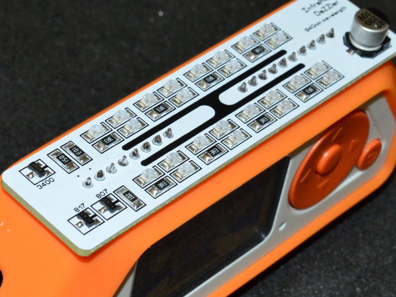 K&ouml;nnte beinhalten: Ein orange-wei&szlig;es elektronisches Ger&auml;t mit einer Leiterplatte mit zahlreichen kleinen Lichtern und Komponenten. Die Leiterplatte tr&auml;gt den Text "Infrared Dazzler" und "940nm wavelength". Das Ger&auml;t hat zwei orangefarbene Kn&ouml;pfe und eine silberne Komponente.
