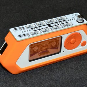 K&ouml;nnte beinhalten: Ein orange-wei&szlig;es elektronisches Ger&auml;t mit einem Bildschirm, der eine Grafik anzeigt. Es hat eine Leiterplatte oben mit LED-Leuchten und dem Text "Intrafied Gezza". Das Ger&auml;t hat ein orangefarbenes Bedienfeld mit einer kreisf&ouml;rmigen Taste.