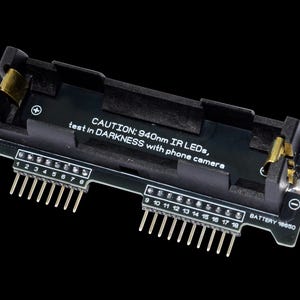 Może przedstawiać: Czarny element elektroniczny o prostokątnym kształcie, z wieloma pinami i komorą na baterie. Komponent zawiera tekst "CAUTION: 940nm IR LEDs, test in DARKNESS with phone camera" i "BATTERY 18650".