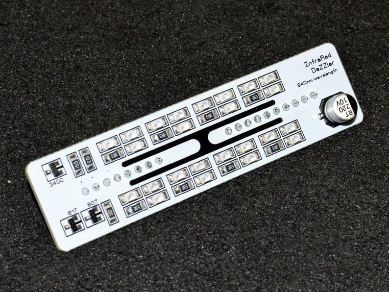 K&ouml;nnte beinhalten: Eine wei&szlig;e Leiterplatte mit der Aufschrift "InfraRed DaZZler" und dem Text "940nm wavelength". Sie verf&uuml;gt &uuml;ber zahlreiche kleine, rechteckige Lichter und ein schwarzes Designelement. Eine kleine Metallkomponente ist ebenfalls vorhanden. Auf dunklem Untergrund.