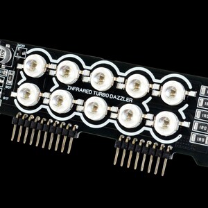 Może przedstawiać: Czarna płytka drukowana z napisem "INFRARED TURBO DAZZLER" i wieloma diodami LED. Płytka posiada piny do podłączenia do innych urządzeń oraz kilka małych element&oacute;w elektronicznych.