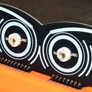 Puede incluir: Componente electr&oacute;nico negro con dos c&iacute;rculos conc&eacute;ntricos blancos y un dise&ntilde;o de l&iacute;nea curva blanca. Dos luces LED est&aacute;n centradas dentro de los c&iacute;rculos. El componente tiene una serie de pines a lo largo del borde inferior. El componente est&aacute; sobre una superficie naranja.