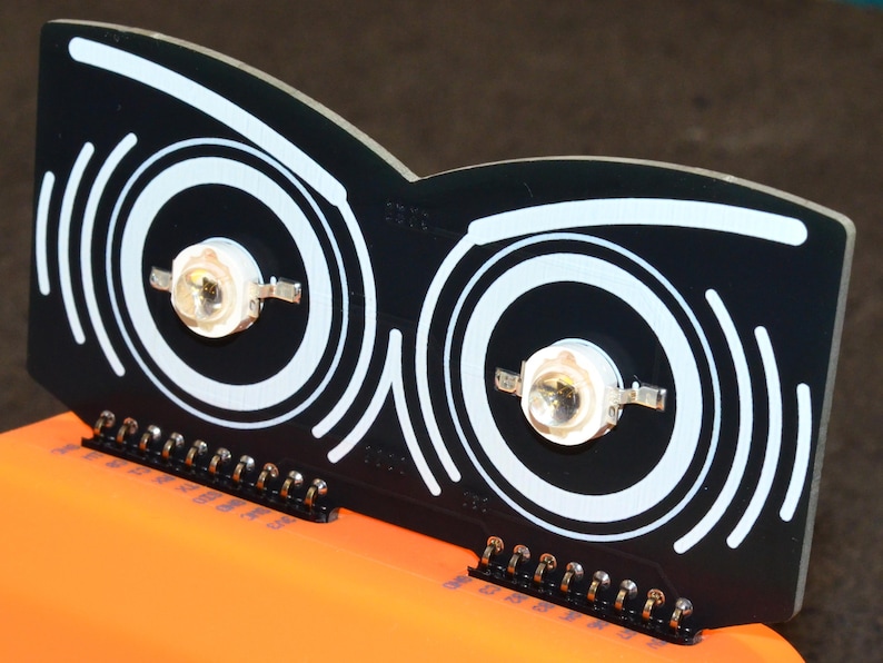 Puede incluir: Componente electr&oacute;nico negro con dos c&iacute;rculos conc&eacute;ntricos blancos y un dise&ntilde;o de l&iacute;nea curva blanca. Dos luces LED est&aacute;n centradas dentro de los c&iacute;rculos. El componente tiene una serie de pines a lo largo del borde inferior. El componente est&aacute; sobre una superficie naranja.