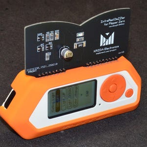 Puede incluir: Un dispositivo electr&oacute;nico naranja y blanco con una placa de circuito negro adjunta. La placa de circuito tiene varios componentes y el texto "InfraRed DaZZler for Flipper Zero". El dispositivo tiene una pantalla que muestra opciones y un bot&oacute;n de control naranja.