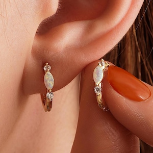 May include: Gold hoop earrings with an oval opal and two clear round stones. The earrings are small and delicate, with a polished finish. The earrings are shown on an ear.