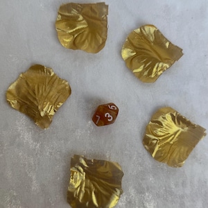 Può includere: Cinque petali di tessuto dorato e strutturato sono disposti attorno a un piccolo dado traslucido arancione-marrone a più facce. Il dado mostra i numeri 1, 3, 5 e 7.