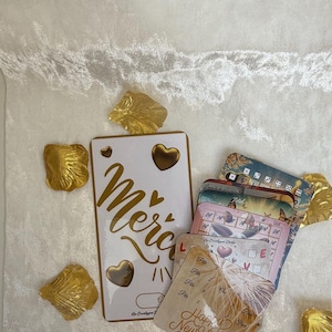 Peut inclure: Ensemble de cartes de vœux et d'éléments décoratifs. Les cartes présentent des lettres dorées et des motifs de cœurs, avec le mot « Merci » en évidence. Des accents dorés en forme de feuilles entourent les cartes, sur fond blanc.