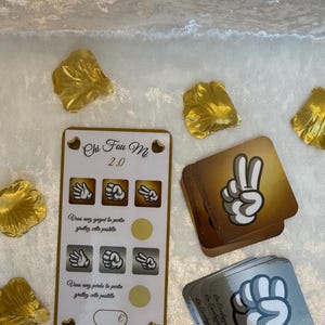 Peut inclure: Un ensemble de cartes de jeu et de décorations en forme de pétales dorés. Les cartes présentent des gestes de la main pour un jeu appelé "Chi Fou Mi 2.0" avec du texte en français. Les cartes sont dans des tons or et argent.