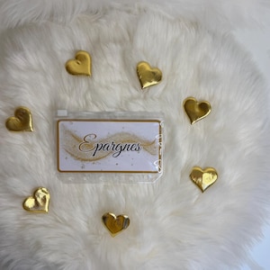 Puede incluir: Una bolsa de plástico transparente con la palabra "Epargnes" impresa, rodeada de decoraciones doradas en forma de corazón. La bolsa y los corazones están sobre una superficie blanca y esponjosa.