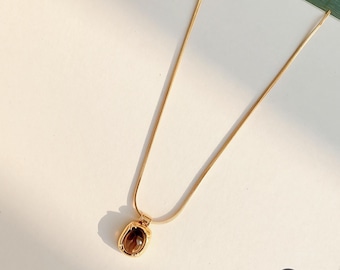 May include: A gold-toned necklace featuring a thin snake chain and a small, rectangular pendant with rounded corners. The pendant contains a smooth, dark brown, oval-shaped stone set within a polished gold-colored metal frame.
