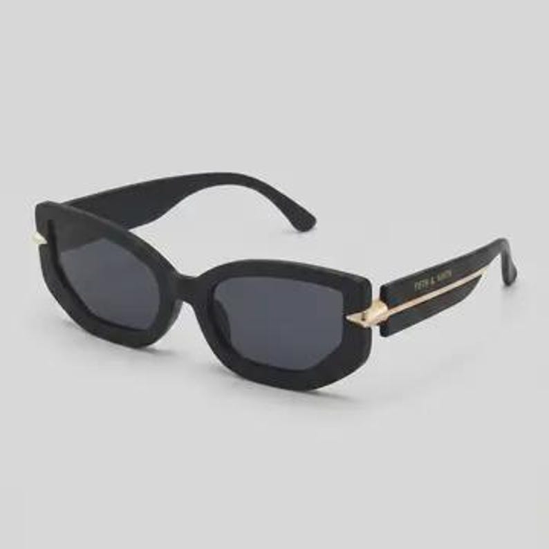 May include: Black sunglasses with a rectangular frame and dark lenses. The sunglasses have gold-colored accents on the temples and the text "FIFTH & NINTH" is printed on the left temple. The sunglasses are set against a light gray background.