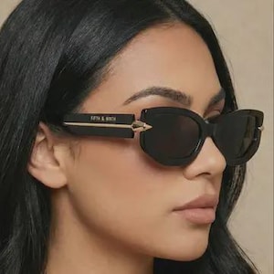 May include: Black sunglasses with a cat-eye shape. The temples feature gold accents and the text "FIFTH & NINTH" in gold lettering. The lenses are dark, and the frames are thick and black.
