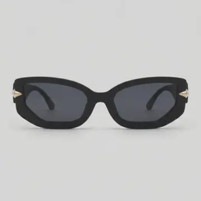 May include: Black sunglasses with a geometric frame and dark lenses. The sunglasses have gold-colored accents on the temples. The overall design is modern and stylish.