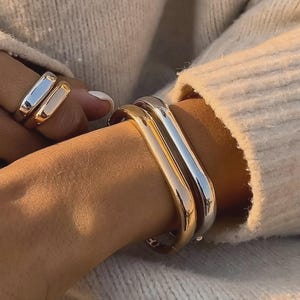 May include: A close-up shot showcases gold and silver-toned jewelry. A gold ring and a silver ring are on a finger. Two matching bracelets, one gold and one silver, are stacked on a wrist. The jewelry is set against a neutral-toned sweater.