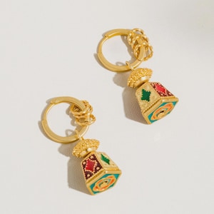 May include: A pair of gold-colored hoop earrings. Each earring features a small, colorful, geometric pendant with red, green, and blue accents. The pendants have a textured, ornate top. The earrings are displayed on a white background.