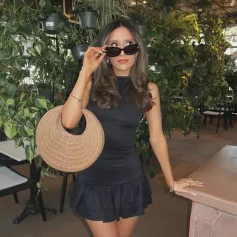 May include: A woman wearing a black dress and sunglasses, holding a round woven handbag. The dress has a high neckline and a ruffled hem. The background features lush greenery and hanging plants.