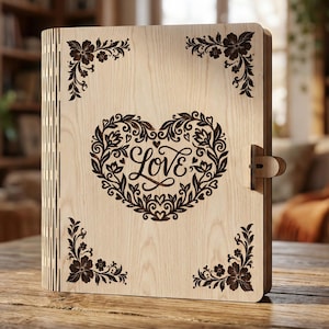 Op de afbeelding: Een houten dagboek met een lasergeëtst ontwerp. De omslag heeft een hartvormige bloemenkrans met het woord "Love" in het midden. Decoratieve bloemenhoekaccenten en een sluiting maken het ontwerp compleet. Het dagboek is gemaakt van lichtgekleurd hout.