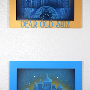 May include: Two framed pictures in blue and gold colours. Both pictures feature a silhouette of a city with a rainbow arch over it. The text "DEAR OLD SHIZ" is at the bottom of each picture.