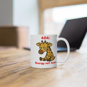 Könnte beinhalten: Weiße Keramik-Tasse mit einem Cartoon-Giraffen-Design und dem Text "404: Energy not found". Die Giraffe ist braun mit gelben Flecken und hat einen traurigen Gesichtsausdruck. Die Tasse steht auf einer Holzoberfläche.