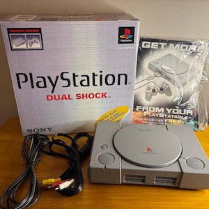 May include: A gray PlayStation console with a Dual Shock box and a "Get More" pamphlet. The box reads "PlayStation Dual Shock" and "Sony". Cables and a yellow sticker are also visible.