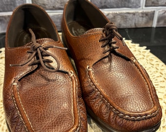 May include: A pair of brown leather shoes with laces. The shoes have a textured surface and white stitching around the edges. The shoes are on a cream-colored, open-weave mat.