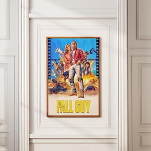 Peut inclure: Une affiche encadrée du film "Fall Guy" avec un homme en veste rouge et une femme avec un mégaphone. L'affiche a un fond bleu avec des scènes d'action et le titre en jaune.