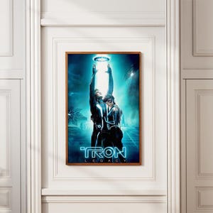 以下が含まれることがあります： 映画「トロン：レガシー」のフレーム入りポスター。アートワークは、未来的な黒と青の衣装を着た2人の人物が、輝く光に向かって手を伸ばしている様子を描いています。「TRON LEGACY」のタイトルは青いネオン文字で表示されています。