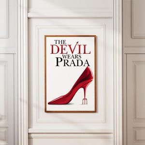 May include: A framed poster with the words "THE DEVIL WEARS PRADA" in red and black. A glossy red high-heeled shoe with a trident heel is on a white background. The poster is in a brown wooden frame, suitable for a fashion-themed room.