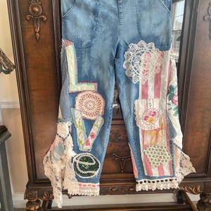 May include: Light blue denim pants with patchwork details. The word "LOVE" is spelled out with fabric and lace appliqués. Patches of floral and striped fabric are sewn onto the pants, along with lace trim.