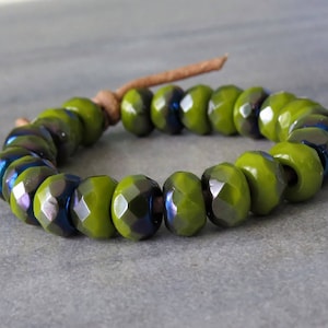 May include: A bracelet featuring faceted olive green beads with iridescent blue and purple undertones. The beads are strung on a brown leather cord, creating a simple yet elegant accessory. The bracelet is shown on a gray surface.