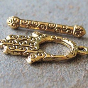 May include: Close-up of two ornate gold-coloured jewellery clasps. One clasp is a bar with decorative swirls, and the other is a hand-shaped toggle clasp. The clasps are likely for necklaces or bracelets, and are made of metal.