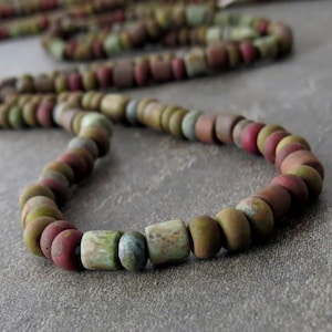 May include: A necklace with a strand of matte, earthy-toned beads. The beads are a mix of round and cylindrical shapes in shades of green, red, brown, and blue. The necklace is displayed on a neutral grey surface.