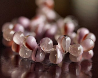 May include: A close-up of a bracelet made of teardrop-shaped beads. The beads are a translucent pink with red and black stripes. The bracelet is resting on a dark brown surface.