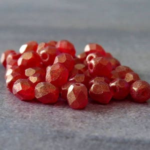 Puede incluir: Un montón de cuentas de vidrio facetadas rojas con un acabado metálico dorado. Las cuentas son pequeñas y tienen una forma ligeramente irregular.