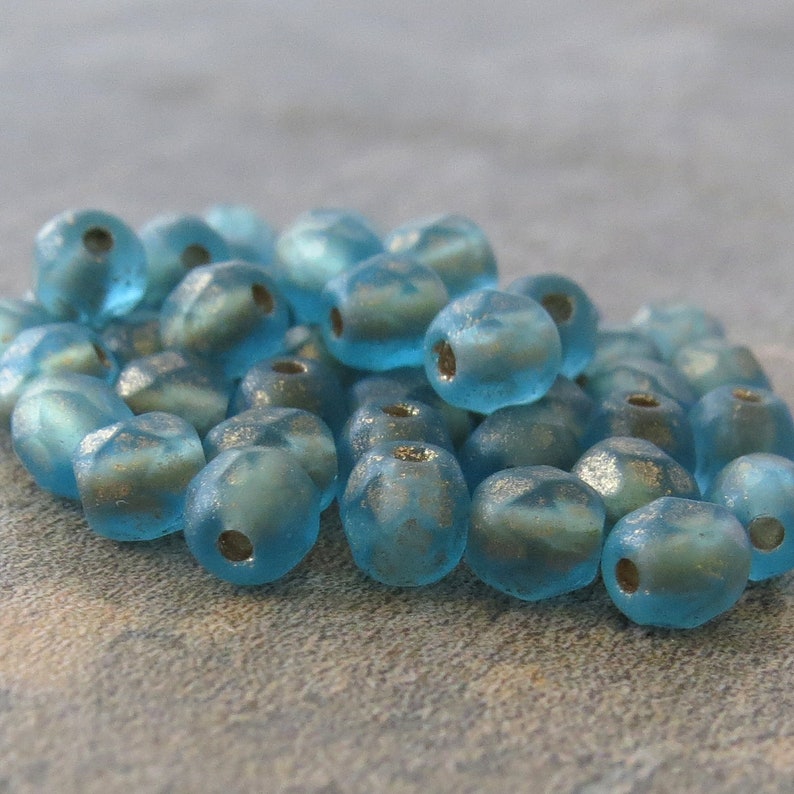 May include: A collection of light blue glass beads with a frosted appearance. Each bead has a rounded shape with facets and a hole for stringing. The beads have a subtle metallic shimmer, suggesting a vintage or handmade quality.