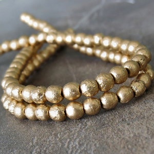 May include: A strand of gold-colored beads with a textured surface. The beads are round and have a matte finish.