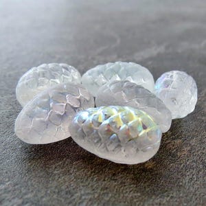 May include: Six iridescent white glass pinecone shaped ornaments. The ornaments have a textured surface that resembles scales.