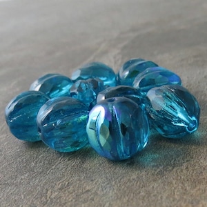 May include: A group of teal blue glass beads with a faceted, iridescent finish. The beads are oval shaped and have a textured surface.