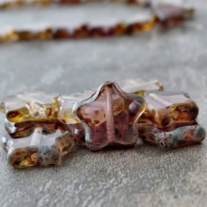 May include: A close-up of a group of star-shaped glass beads. The beads are a mix of purple, brown, and gold colours. The beads are translucent and have a shimmery finish.