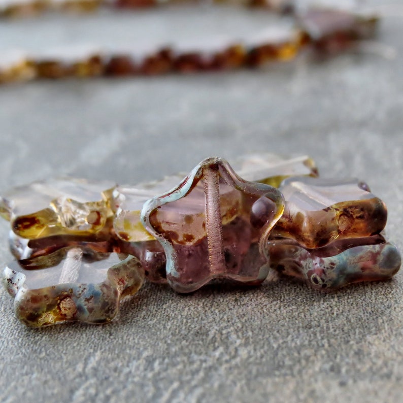 May include: A close-up of a group of star-shaped glass beads. The beads are a mix of purple, brown, and gold colours. The beads are translucent and have a shimmery finish.