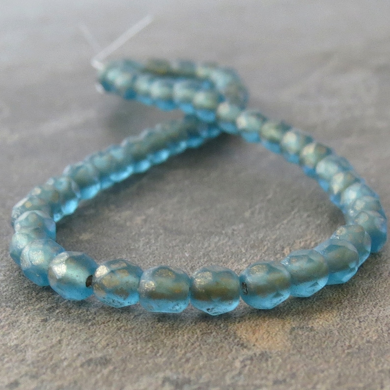 May include: A strand of faceted, light blue glass beads. The beads have a slightly irregular shape and a subtle, iridescent sheen. The beads are strung on a thin, clear cord, suitable for jewelry making or craft projects.