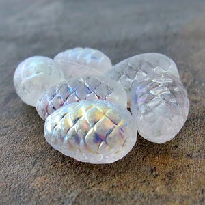 May include: A group of six iridescent white glass pine cone shaped beads. The beads have a textured surface and a pearlescent finish.