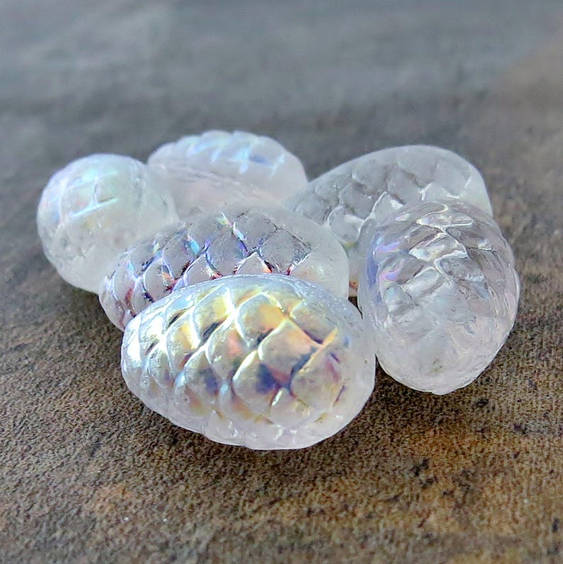 May include: A group of six iridescent white glass pine cone shaped beads. The beads have a textured surface and a pearlescent finish.