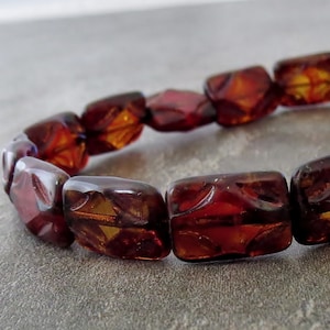 May include: A close-up of a dark red amber bracelet with square-shaped beads. The beads have a carved design and are strung together on a clear elastic cord.
