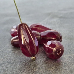 May include: A collection of deep red, faceted glass beads with metallic bronze-colored vertical accents. One bead is suspended on a thin gold-toned wire, while the others are clustered together, showcasing their teardrop shape and reflective surfaces.