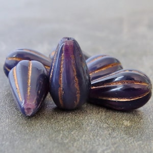 May include: A group of purple glass beads with gold accents. The beads are shaped like teardrops and have a ribbed texture.